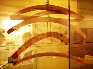 In einer Glasvitrine hängen verschiedene, hölzerne Kleiderbügel. Eines davon trägt die Aufschrift "Kaufhaus zur billigen Quelle L. Reches Marburger Tor 4 & 24"