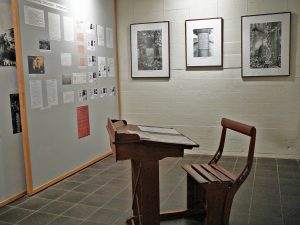 Ein altes, hölzernes Schreipult mit Sitzbank im Vordergrund, Ausstellungstafeln im Hintergrund.