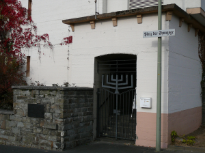 Geschlossenes, geschmiedetes Eingangstor, dessen oberer Teil wie eine jüdische Menora geformt ist. Neben dem Eingang ein Straßenschild mit dem Aufdruck "Platz der Synagoge".