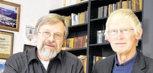 Zwei ältere Männer mit Brille blicken in die Kamera. Im Hintergrund ein Bücherregal.